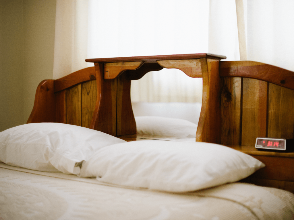 A photograph by Alec McClay. A wooden bed frame, a made bed with two white pillows and white sheets. An analog clock that reads 3:14.